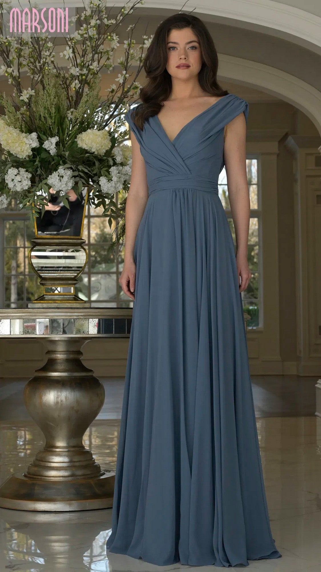 A woman wearing the Marsoni M251 long formal dress stands indoors near a large floral arrangement and elegant décor.